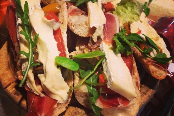 Open sandwiches with cheese, cured meat, and arugula on wooden platter.