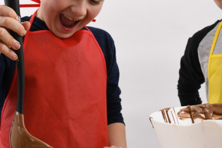 Two children making chocolates with ladle and molds, wearing colorful aprons.