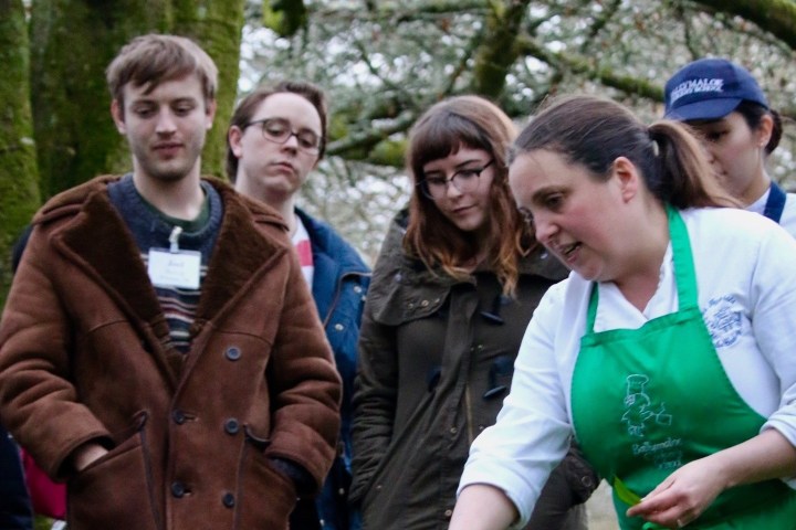 A group of people outdoors, with a woman wearing a green apron demonstrating something.