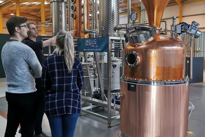 Three people observing a large copper distillation apparatus inside an industrial building.