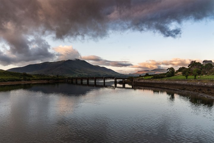 Mountain landscape with a river, bridge, and cloudy sky at sunset.