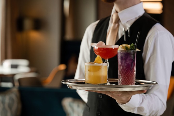 A waiter holding a tray with three colorful cocktails in a dimly lit restaurant.