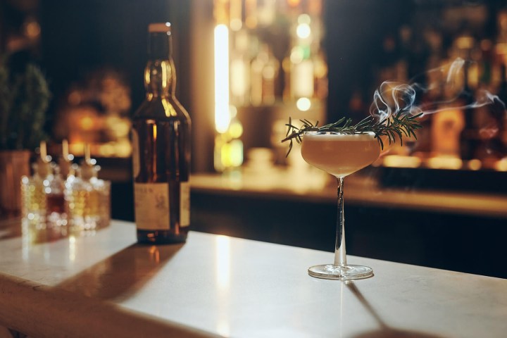 Cocktail on bar with rosemary garnish, smoke, and a bottle in the background.