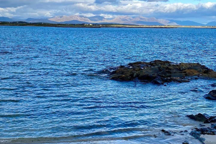 Rocky shoreline with blue sea and mountains under a partly cloudy sky.