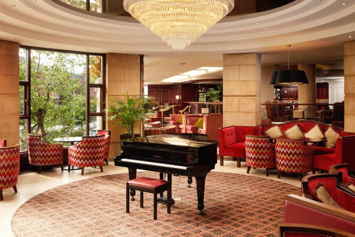 Elegant lounge with grand piano, red chairs, large windows, and chandelier.