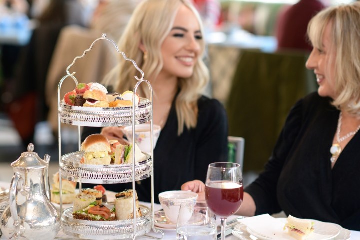 Two women enjoying afternoon tea with a three-tier tray of snacks and drinks.