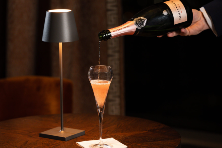 Person pouring pink champagne into a glass next to a small table lamp.