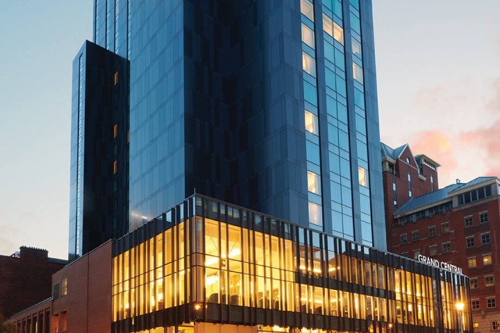 Tall hotel building with glass facade lit from within, labeled Grand Central, against evening sky.