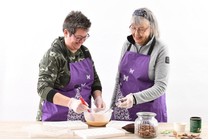Two women in purple aprons mixing ingredients in a bowl, with molds and jars on the table.