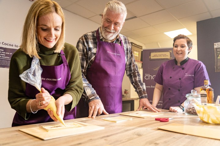Three people in aprons decorating chocolate on wooden boards in a workshop.