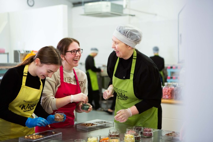 Three women in aprons laughing while making chocolates in a kitchen.