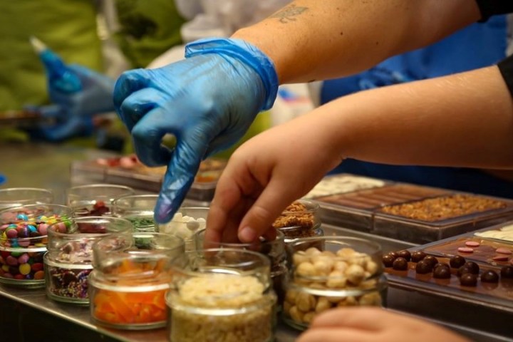 Hands selecting toppings from jars for chocolate bars, wearing blue gloves.