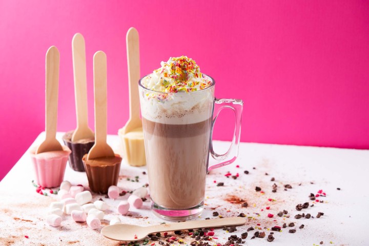 Hot chocolate with whipped cream and sprinkles, surrounded by marshmallows and cocoa sticks.
