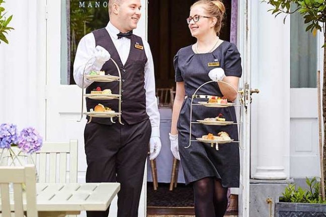 Two servers carrying trays with desserts walk outside a building labeled 'Hayfield Manor.'