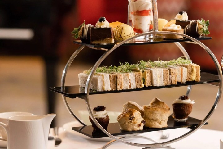 Three-tiered afternoon tea stand with desserts, sandwiches, and scones on a table with a teapot.