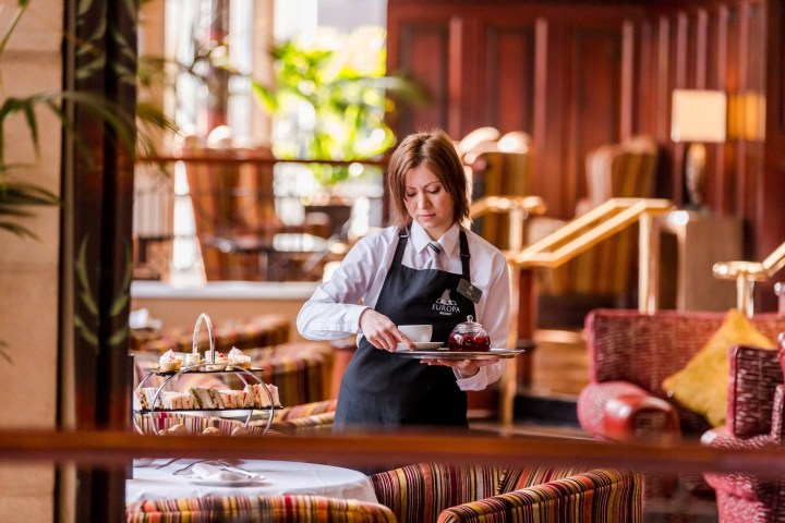 Waitress serving tea on a tray in a hotel lounge with colorful chairs and tables.