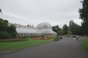 Victorian glasshouse in a park with flower beds and a path, overcast sky.