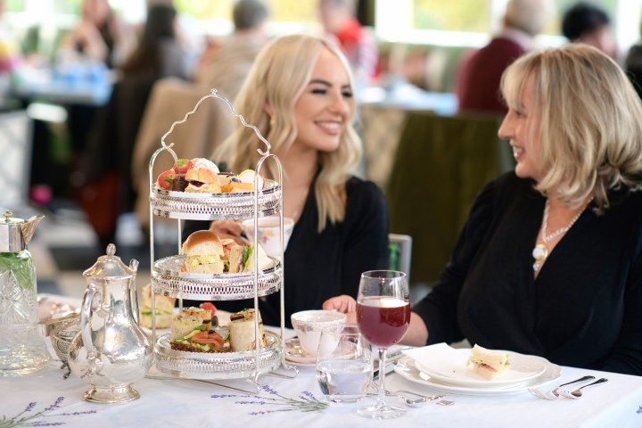 Two women enjoy afternoon tea with a tiered tray of snacks and drinks on a table.