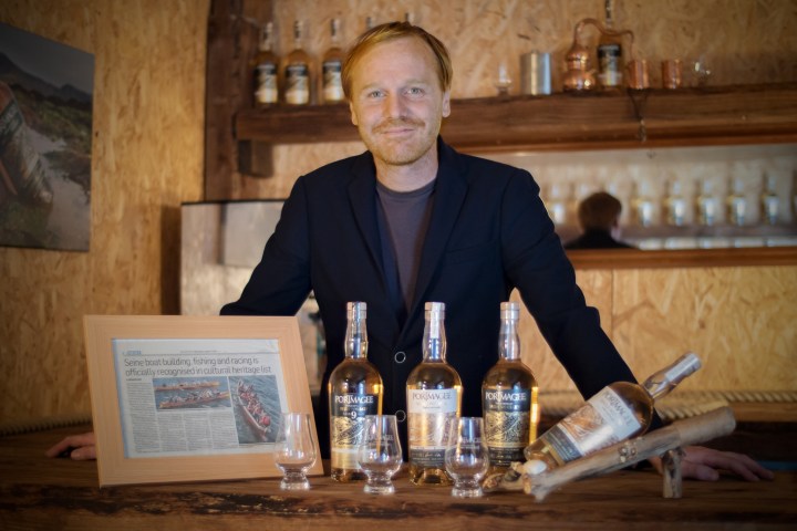 Man smiling behind whiskey bottles and framed article on a wooden counter.