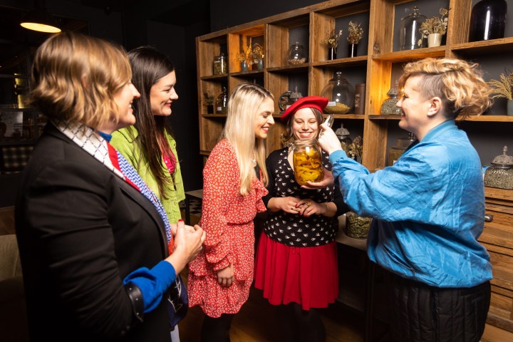 Five women in a room with wooden shelves and jars, smiling and holding a jar.