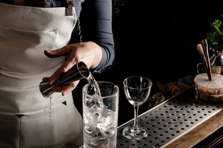Bartender pouring liquid into a glass with ice on a bar counter.