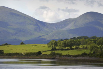 Scenic view of lush green hills, trees, and a calm river under a partly cloudy sky.