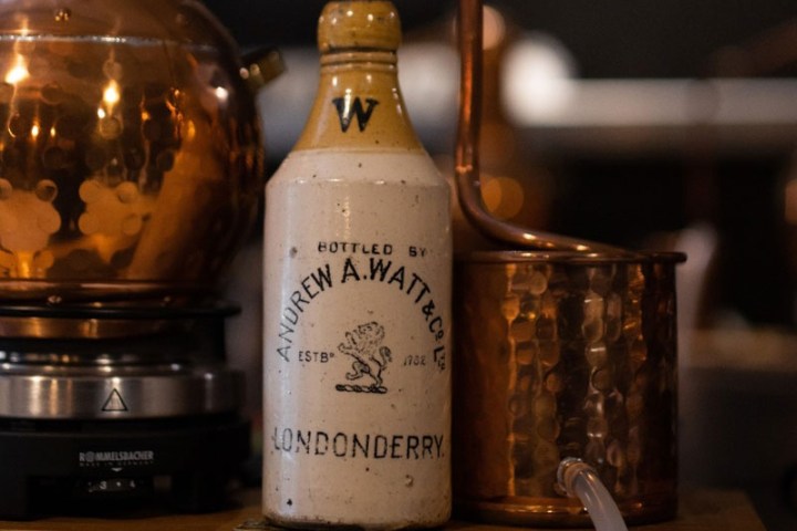 Ceramic bottle labeled 'Andrew A. Watt & Co Ltd Londonderry' next to copper distilling equipment.