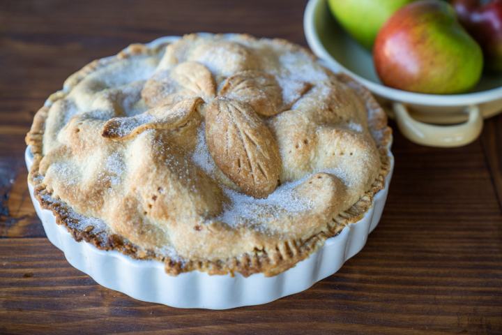 A freshly baked apple pie in a white dish, with apples in a bowl nearby.