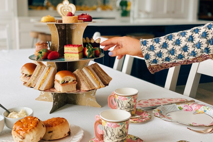 Hand reaching for pastries on a tiered stand with sandwiches, scones, and tea cups on a table.