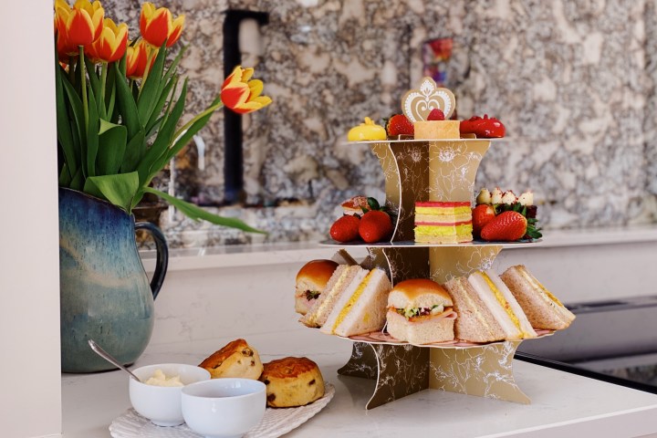 Three-tiered cake stand with sandwiches and desserts beside tulips and scones on a kitchen counter.