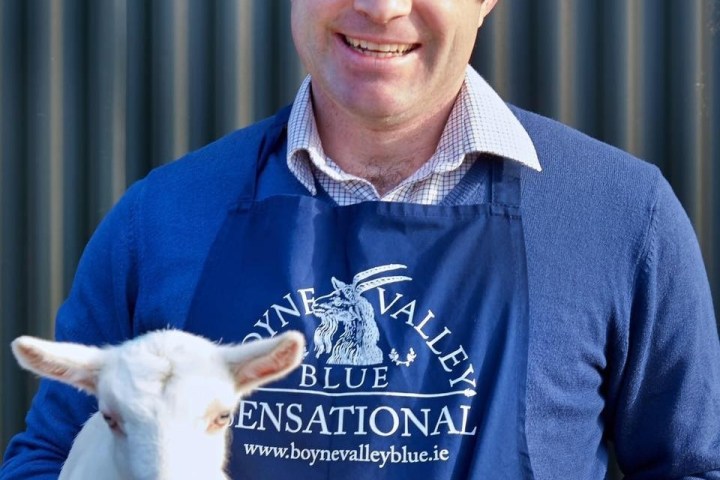 Man in blue apron holds a small white goat and cheese block, smiling.