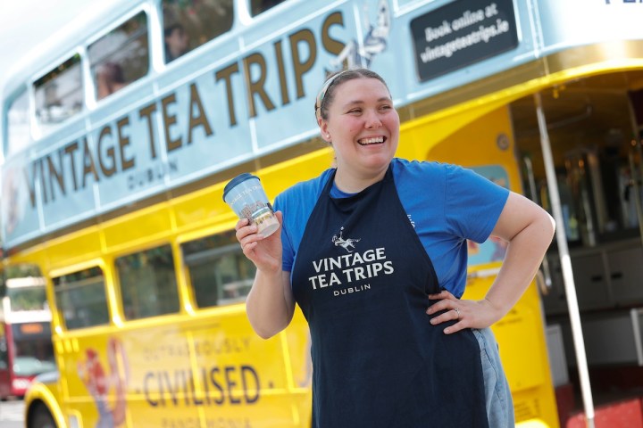 Person smiling, holding a cup in front of a yellow vintage tea bus.