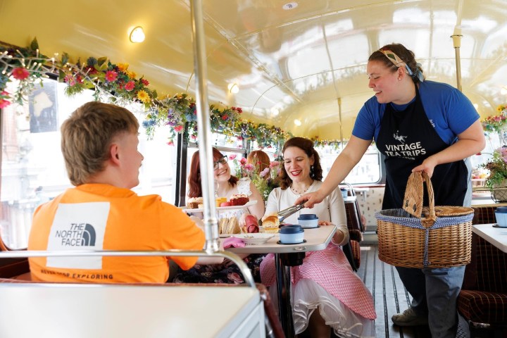 People enjoying tea on a vintage bus, served by a smiling person in a blue shirt and apron.