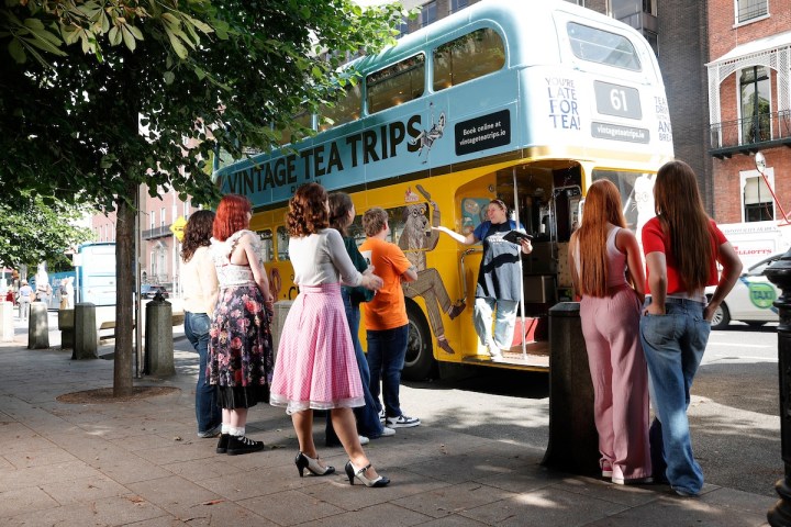People queue outside a vintage tea bus named 'Vintage Tea Trips' on a city street.