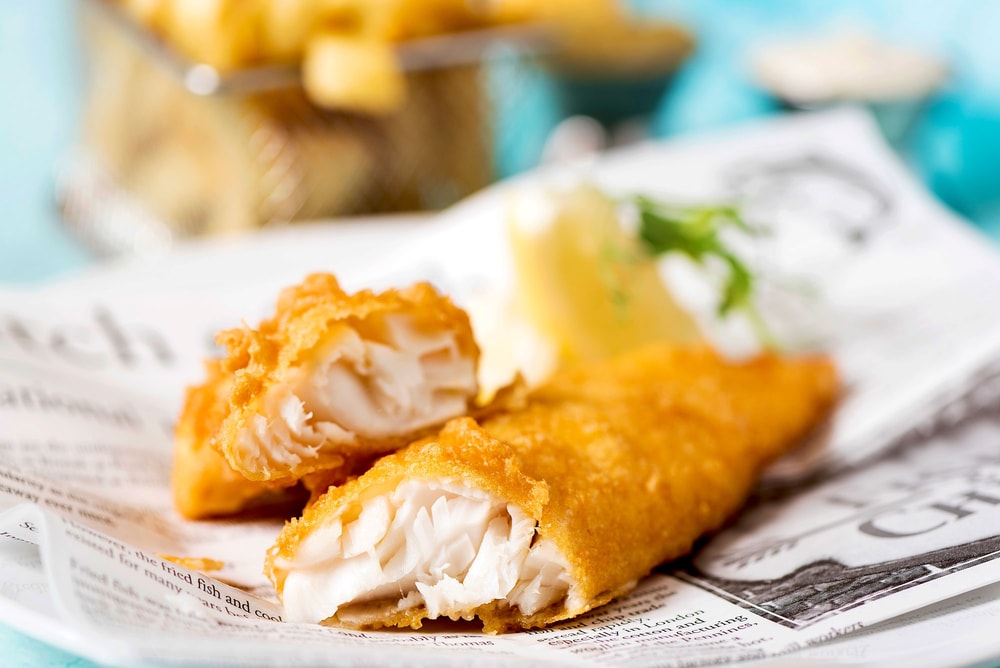 Close-up of battered fish on newspaper, with fries in the background.