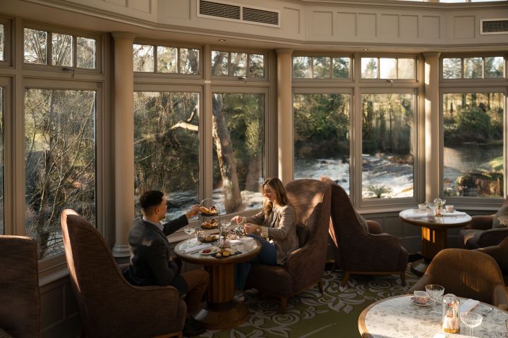 Couple enjoying afternoon tea in a sunlit room with large windows view of river and trees.