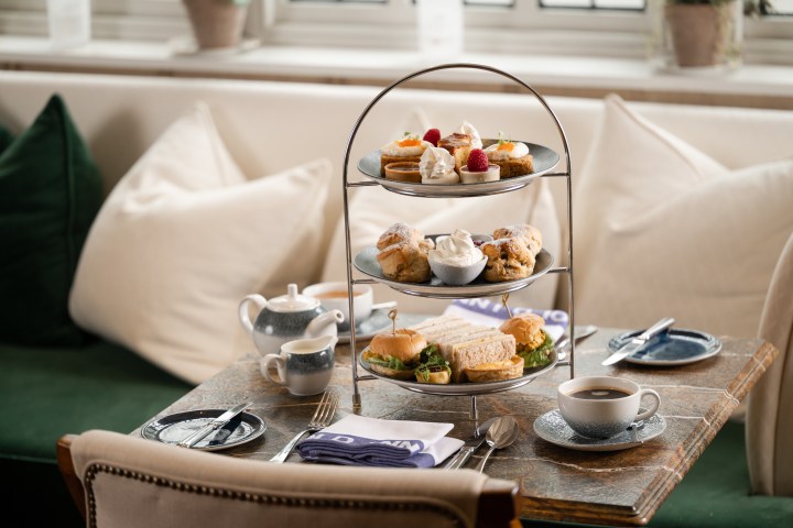 Three-tiered tray with sandwiches, scones, and pastries on a table with tea set and napkins.