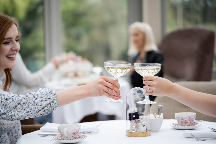 Two women clinking glasses in a bright restaurant, smiling with tea cups on the table.