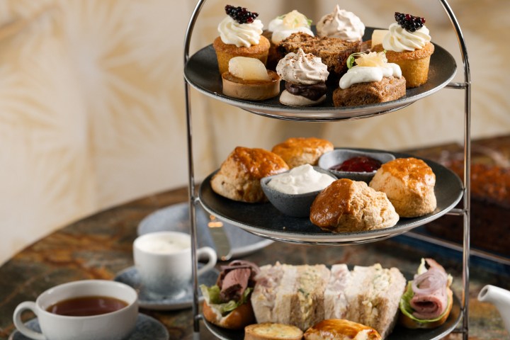 Three-tiered afternoon tea stand with pastries, scones, sandwiches, and two cups of tea on a marbled table.