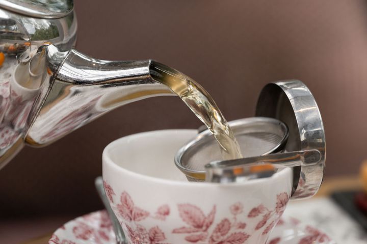 Tea being poured from a silver teapot into a floral teacup with a strainer.