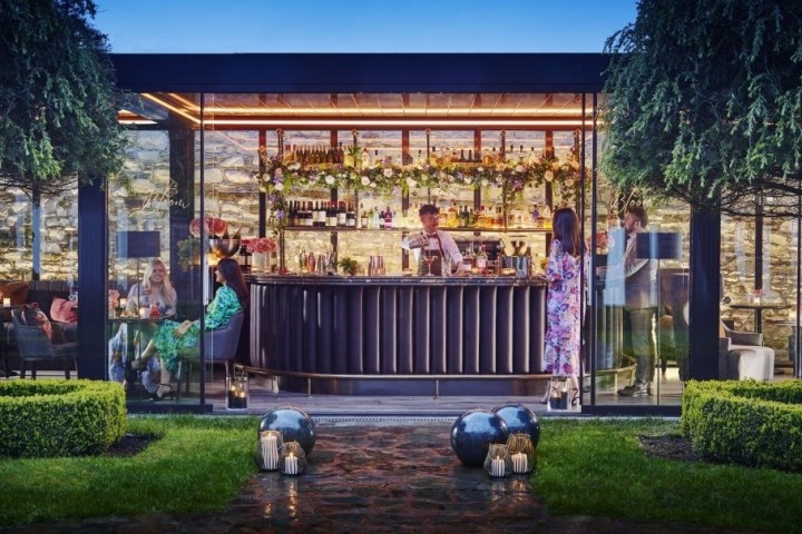 Outdoor glass-walled bar with people inside, surrounded by trees and greenery.
