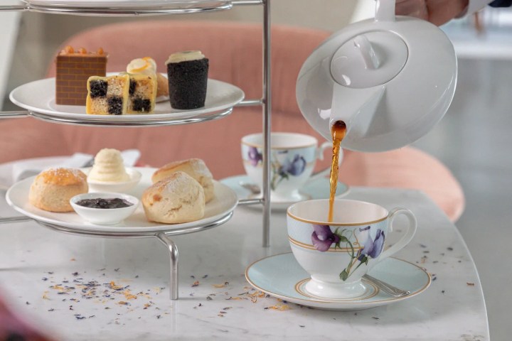 Tea being poured into floral cup by a tiered tray with scones and pastries on a marble table.