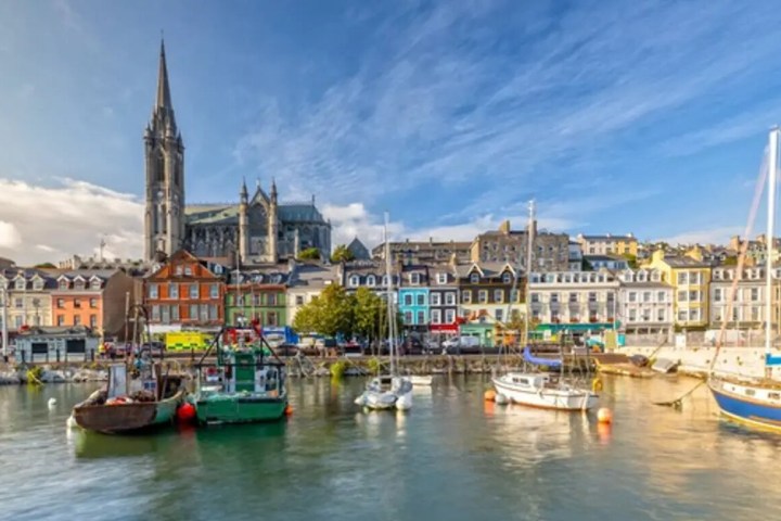 Colorful buildings and boats by a waterfront under a clear sky, with a tall church spire in the background.