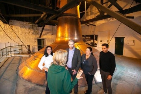 Five people in discussion near a large copper still in a distillery.