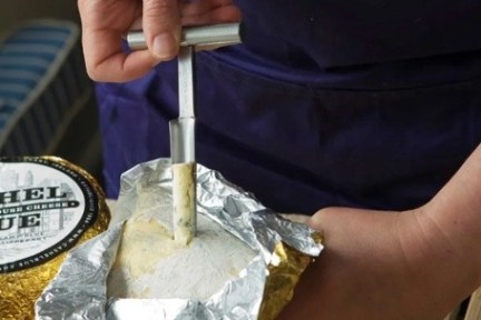 Person holding cheese sample tool over wrapped cheese wheel.