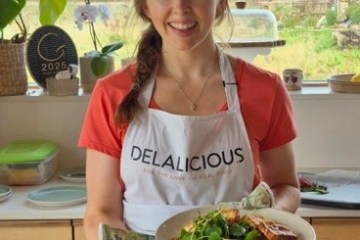 Woman in kitchen holding two plates of food, wearing a 'Delalicious' apron.