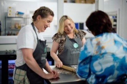 Three women smiling and talking while one rolls dough in a kitchen.