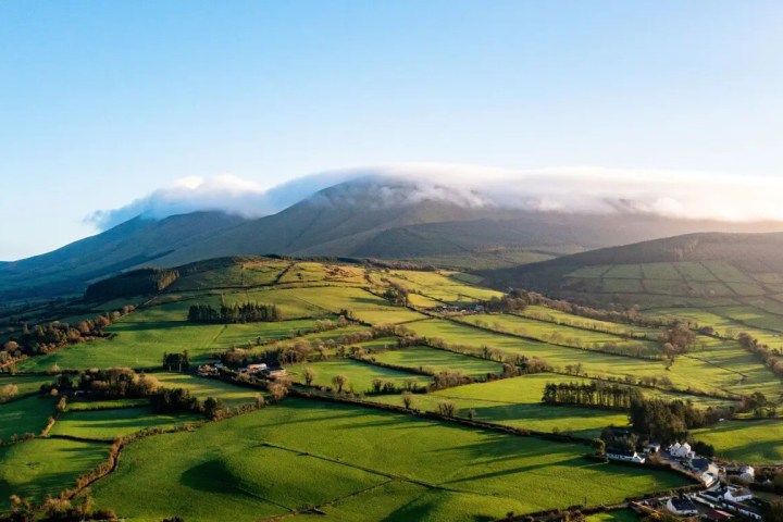 Lush green fields with distant mountains under a blue sky and cloud cover.
